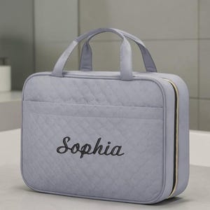 May include: A light gray rectangular travel bag with a quilted diamond pattern and two top handles. The front features the name Sophia embroidered in black script. The bag includes a side zipper closure with gold-toned hardware and a front pocket.