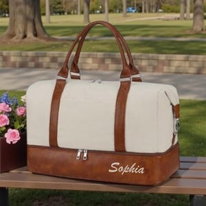 May include: Cream-colored duffel bag with brown leather accents and handles. The bottom section and straps are brown leather. The bag has a zipper closure and the name "Sophia" is embroidered on the front. The bag is sitting on a wooden bench.
