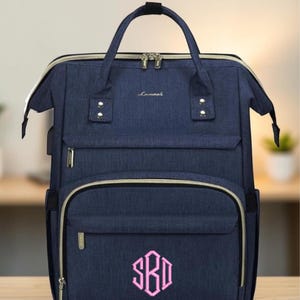 May include: A navy blue backpack with a textured fabric finish, featuring a top handle, dual shoulder straps, and multiple zippered compartments. The front pocket displays a pink embroidered monogram with the letters SBD in a hexagonal frame.