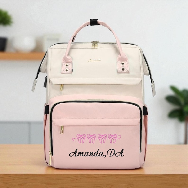 May include: A cream and pink backpack with a top handle and shoulder straps. The front pocket features a pink embroidered design of teeth and the text "Amanda, D.A." The bag has gold zippers and black accents.