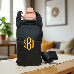May include: A black quilted water bottle carrier with an adjustable strap and a zippered front pocket. The front features an embroidered yellow monogram with the letters SBD in a hexagonal frame. A light pink water bottle is inserted into the top.