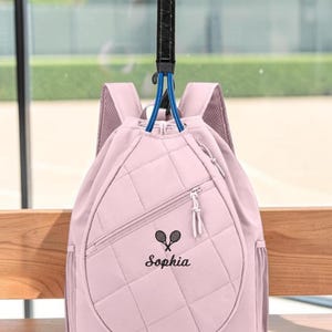 May include: A light pink quilted backpack featuring a diagonal zippered pocket, side mesh pockets, and adjustable shoulder straps. The front displays a black embroidered graphic of two crossed tennis rackets above the name Sophia in script font.