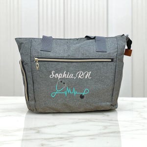 May include: A heather gray tote bag featuring a horizontal zippered pocket, two top handles, and white embroidered text that reads Sophia, RN. Below the name is a teal embroidered graphic of a stethoscope forming a heartbeat rhythm line.