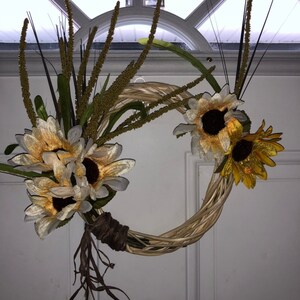 May include: A brown wicker wreath decorated with yellow and white artificial sunflowers. The wreath is hanging on a white door.