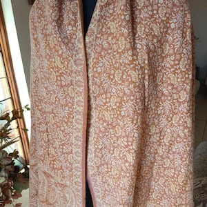 Könnte beinhalten: Ein pfirsichfarbener Schal mit einem komplizierten Blumen- und Paisley-Muster. Der Schal hat einen Fransenrand und ist über einer Schaufensterpuppe drapiert. Das Design weist weiße und gelbe Akzente auf und schafft ein detailliertes und elegantes Textil.