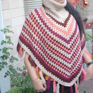 Puede incluir: Un poncho de ganchillo hecho a mano con un patrón geométrico, con tonos beige, rosa y marrón. El poncho tiene un borde con flecos y un cuello alto beige a juego. Una prenda cómoda y elegante para el clima fresco.