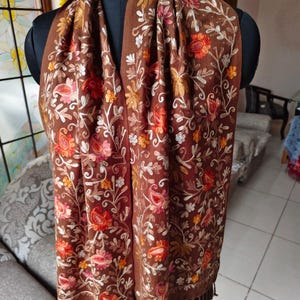 May include: A brown scarf with intricate floral embroidery in shades of red, orange, pink, and white. The scarf is draped over a mannequin, showcasing the detailed pattern and fringed edges. The design features paisley and floral motifs.