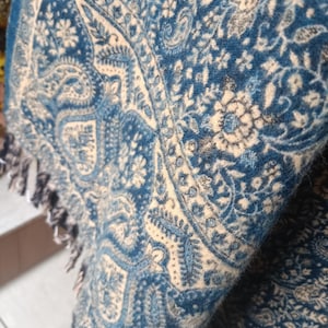 Puede incluir: Primer plano de un chal azul y crema con un intrincado estampado de cachemira. El chal tiene un aspecto suave y texturizado y está rematado con flecos en el borde. El diseño presenta elementos florales y curvos.