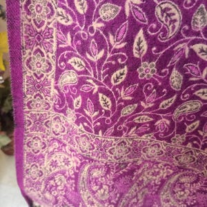 Puede incluir: Primer plano de un chal morado con un estampado floral y cachemir. El chal tiene un diseño color crema con hojas y flores. El borde tiene un patrón floral detallado. El chal está hecho de un material suave y tiene borlas.