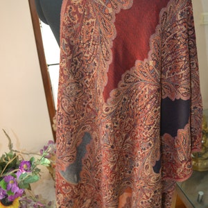 May include: A shawl with a paisley design in shades of red, orange, and brown. The shawl is draped over a mannequin, showcasing the intricate pattern and color palette. The edges are finished with a decorative border.