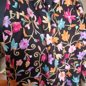 May include: Black scarf with colorful floral embroidery. The scarf features a detailed pattern of pink, purple, orange, and blue flowers with gold-colored stems and leaves. The embroidery is set against a black background.