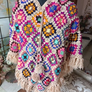 Puede incluir: Un poncho de crochet colorido con flecos y pompones. El poncho está hecho de una variedad de colores, incluyendo rosa, azul, verde, amarillo y naranja. El poncho está en un maniquí.