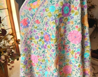 May include: A beige shawl with intricate floral embroidery in shades of pink, purple, blue, green, and orange. The shawl is draped over a mannequin, showcasing the detailed craftsmanship and fringed edges. The design features a repeating pattern of flowers and leaves.
