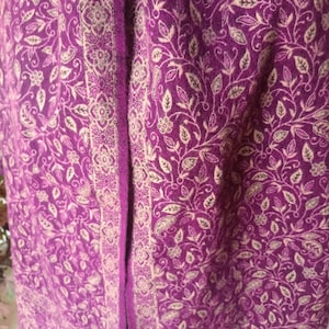 May include: A purple shawl featuring an intricate cream-colored floral and paisley pattern throughout the fabric. The garment includes a distinct vertical border with a repeating geometric and floral design that runs along the edge of the textile.