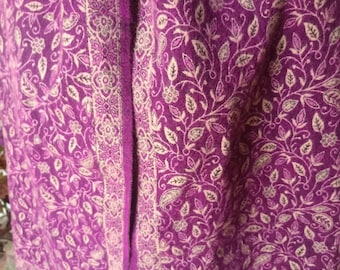 May include: A purple shawl featuring an intricate cream-colored floral and paisley pattern throughout the fabric. The garment includes a distinct vertical border with a repeating geometric and floral design that runs along the edge of the textile.