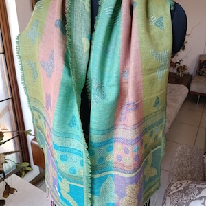 May include: A patterned scarf in pastel shades of pink, green, and blue, adorned with butterfly motifs. The scarf is displayed on a mannequin. The design includes decorative borders and fringed edges. The scarf is a blend of colours and patterns.