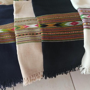 Può includere: Una collezione di scialli piegati in vari colori, tra cui blu navy, nero e crema. Ogni scialle presenta una fascia decorativa con un motivo geometrico nei toni del rosso, verde e giallo. I bordi sono rifiniti con frange.