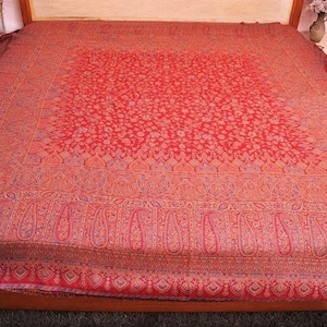 Könnte beinhalten: Eine große rechteckige Tagesdecke mit einer leuchtend roten Grundfarbe und komplizierten, sich wiederholenden Blumen- und Paisley-Mustern in Gold-, Orange- und Blautönen. Das Design umfasst ein zentrales quadratisches Feld mit einer breiten Bordüre.