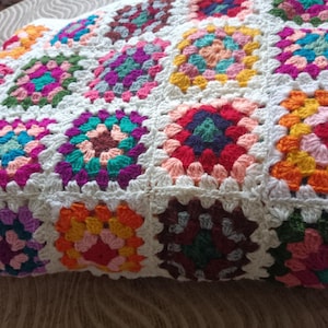 May include: A colorful, handmade crochet pillow with a granny square design. The pillow features squares in various colors, including pink, orange, green, and purple, all set against a white background. The crochet stitches are clearly visible, showcasing the handcrafted nature of the item.