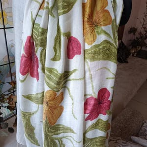 May include: White scarf with a floral pattern featuring red, yellow, and green flowers. The scarf is draped, showcasing the design and texture of the fabric. The flowers are painted with detail.