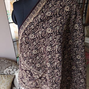 May include: A dark brown shawl featuring an intricate, dense pattern of cream-colored floral motifs and paisley shapes. The fabric displays a consistent, repeating design across the entire surface, finished with a wide, decorative border.