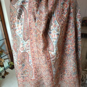 May include: A beige shawl with a paisley pattern in shades of brown, red, and blue. The shawl has a fringed edge and is made of a soft, lightweight fabric.