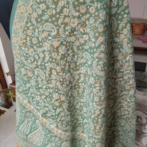 May include: A green shawl featuring a detailed floral and paisley design. The shawl has a fringe along the bottom edge and is draped over a mannequin. The color palette includes shades of green, beige, and light blue.