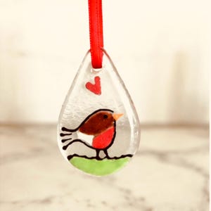 May include: A teardrop-shaped clear glass ornament featuring a painted robin bird with a brown back, red chest, and orange beak. The bird stands on a green patch, with a small red heart above it. A bright red ribbon is threaded through the top hole.