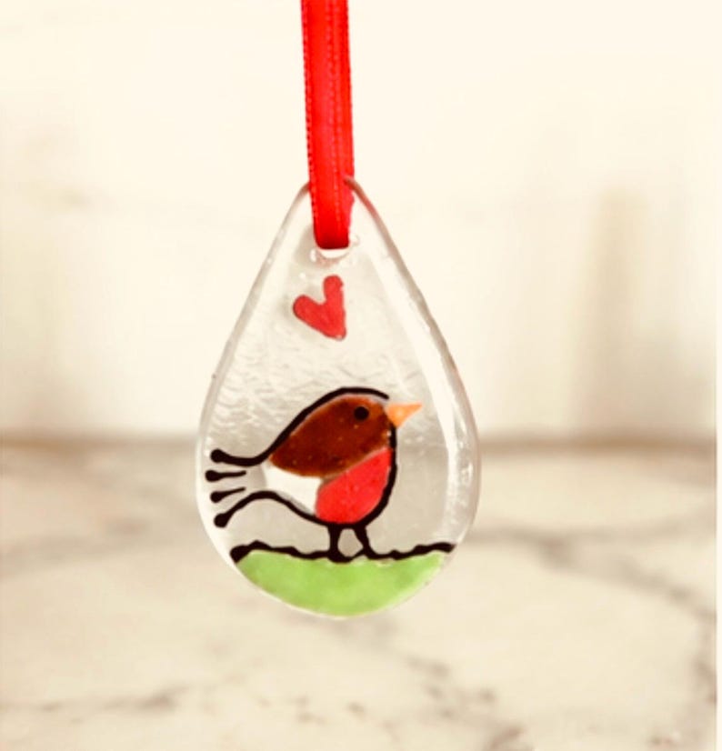 May include: A teardrop-shaped clear glass ornament featuring a painted robin bird with a brown back, red chest, and orange beak. The bird stands on a green patch, with a small red heart above it. A bright red ribbon is threaded through the top hole.