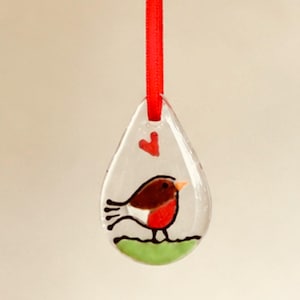 May include: A clear, teardrop-shaped glass ornament featuring a painted robin bird with a brown back, red breast, and orange beak standing on a green patch of grass. A small red heart is painted above the bird, and a red ribbon is attached to the top.