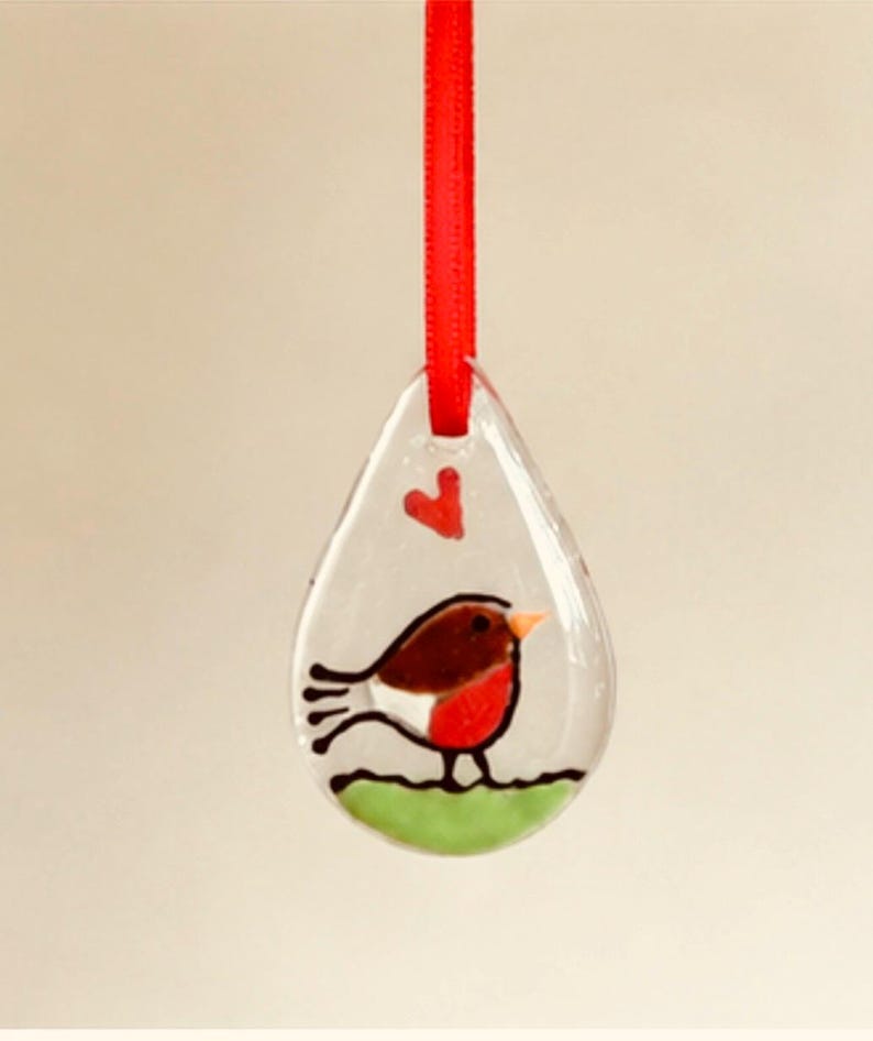 May include: A clear, teardrop-shaped glass ornament featuring a painted robin bird with a brown back, red breast, and orange beak standing on a green patch of grass. A small red heart is painted above the bird, and a red ribbon is attached to the top.