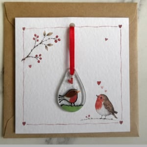 May include: A square greeting card with a brown envelope featuring a teardrop-shaped glass ornament hanging from a red ribbon. The card displays a watercolour illustration of a robin on the ground and a branch with red berries, accented by small red hearts.