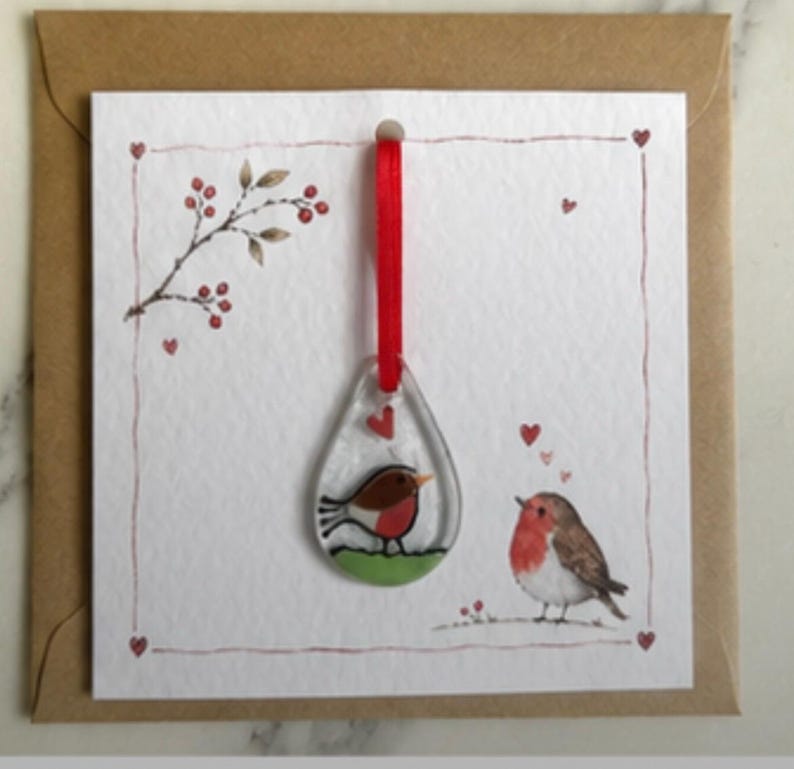 May include: A square greeting card with a brown envelope featuring a teardrop-shaped glass ornament hanging from a red ribbon. The card displays a watercolour illustration of a robin on the ground and a branch with red berries, accented by small red hearts.