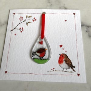 May include: A teardrop-shaped clear glass ornament featuring a painted robin on a green patch, suspended by a red ribbon on a white card. The card displays a matching illustration of a robin, red hearts, and a branch with red berries.