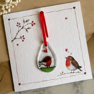 May include: A white square greeting card featuring a red-bordered frame with small red hearts and a branch with red berries. A teardrop-shaped glass ornament with a red ribbon depicts a robin on green grass, accompanied by a robin illustration.