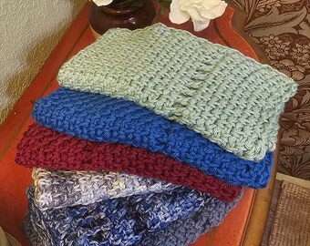 May include: A stack of five crocheted dishcloths in various colors, including green, blue, red, and gray. The dishcloths are folded and stacked on a wooden table with a red surface.