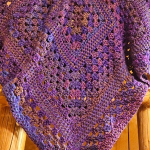 May include: A purple, blue, and brown crocheted throw blanket with a square pattern. The blanket is draped over two wooden posts.