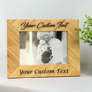 May include: A natural bamboo picture frame with a black engraved inscription "Your Custom Text" on the top and bottom. The frame is holding a black and white photo of flowers in a vase. The frame is 5x7 inches and is sitting on a white surface.