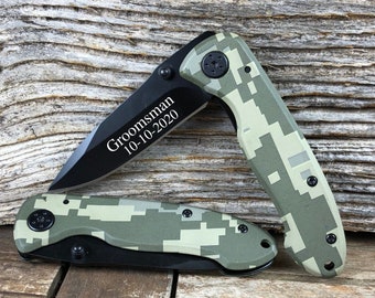 May include: A black pocket knife with a camouflage pattern. The knife has a blade and a handle. The handle is engraved with the text "Groomsman 10-10-2020".