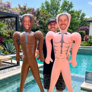 Könnte beinhalten: Zwei aufblasbare Figuren, eine braune und eine rosa, jeweils mit einem lächelnden Gesicht und einem muskulösen Torso-Design. Die Figuren werden von einer Person in der Nähe eines Swimmingpools gehalten. Der Hintergrund umfasst ein Haus, Bäume und einen sonnigen Himmel.