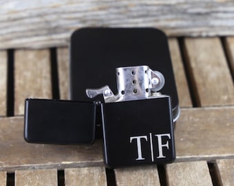 May include: Black lighter with a personalized engraving of the letters "T|F" on the front. The lighter is open and the flint wheel is visible.