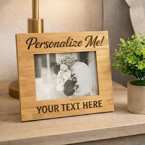 May include: A light brown bamboo picture frame with the text "Personalize Me!" and "YOUR TEXT HERE" engraved on it. The frame holds a black and white photo. A small potted plant sits next to the frame on a wooden nightstand.