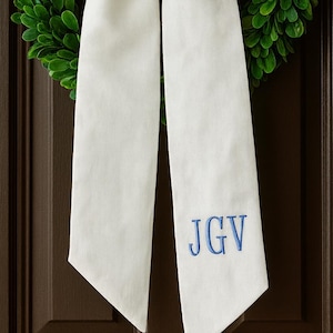 Puede incluir: Una corona de boj verde con un gran lazo blanco y largas cintas. El lazo está centrado en la corona y las cintas cuelgan. Las iniciales bordadas en azul "JGV" están en una cinta. La corona está colgada en una puerta marrón.