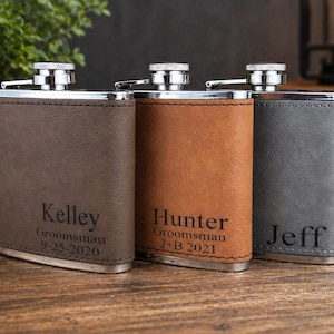 May include: Three personalized hip flasks in brown, tan, and gray leather. Each flask has a silver cap and is engraved with a name and the text "Groomsman" along with a date. The flasks are displayed on a wooden surface.