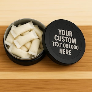 Op de afbeelding: Een open zwarte blik gevuld met witte zakjes, naast een zwart deksel. Het deksel heeft de witte tekst "YOUR CUSTOM TEXT OR LOGO HERE". De blik staat op een houten oppervlak.