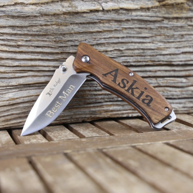 May include: A brown wooden pocket knife with a silver blade. The knife is engraved with the words "Best Man" and "Askia".