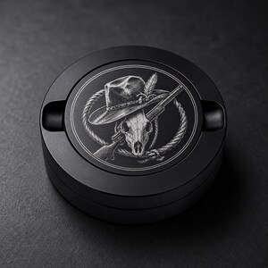 May include: A round black metal ashtray featuring a laser-engraved design on the lid. The graphic depicts a skull wearing a wide-brimmed hat, positioned in front of a rifle and a coiled rope, all rendered in fine white lines against the dark surface.
