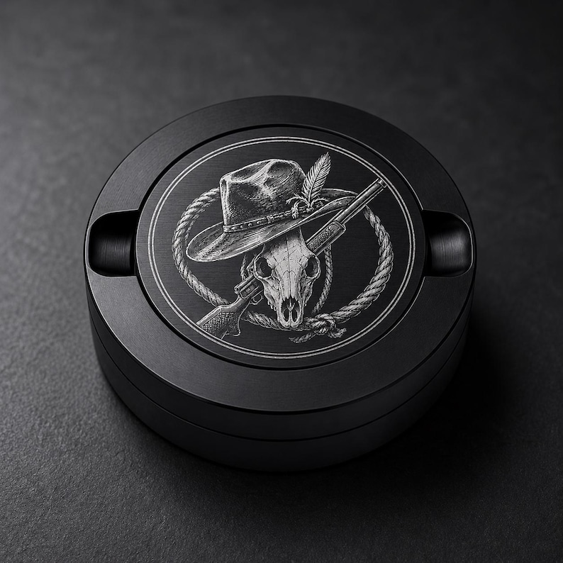 May include: A round black metal ashtray featuring a laser-engraved design on the lid. The graphic depicts a skull wearing a wide-brimmed hat, positioned in front of a rifle and a coiled rope, all rendered in fine white lines against the dark surface.