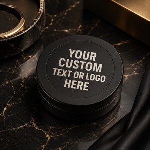 May include: A round, matte black metal tin with a lid featuring the engraved text "YOUR CUSTOM TEXT OR LOGO HERE" in a light, sans-serif font. The container has a smooth, circular design and is presented as a customisable storage vessel.