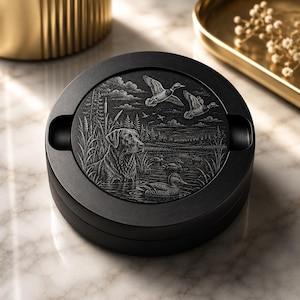 May include: A round black ashtray featuring a detailed monochromatic engraving on its lid. The artwork depicts a Labrador retriever sitting in tall grass near a marsh, with several ducks swimming in the water and two ducks flying overhead.